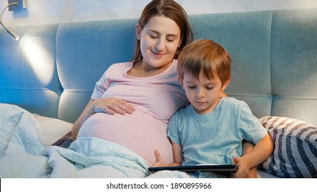 Little Toddler Boy Using Tablet Computer Lying With Pregnant Mother In Bed At Night.