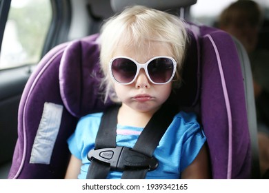 A Little Toddler Child Is Pouting As She Sits Buckled Into The Safety Restraint Harness Of A Car Seat.