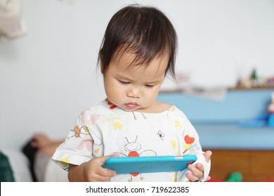 Little Toddler Girl Illness Sitting On Sickbed In Hospital With A Saline Intravenous (IV) On Hand Holding Smartphone And Watching On Youtube