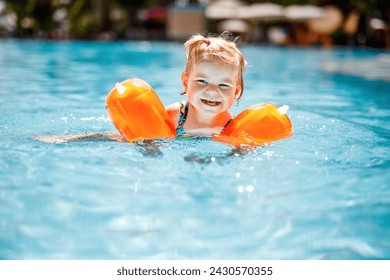 Little toddler girl with protective swimmies playing in outdoor swimming pool by sunset. Baby Child learning to swim in outdoor pool, splashing with water, laughing and having fun. Family vacations. - Powered by Shutterstock