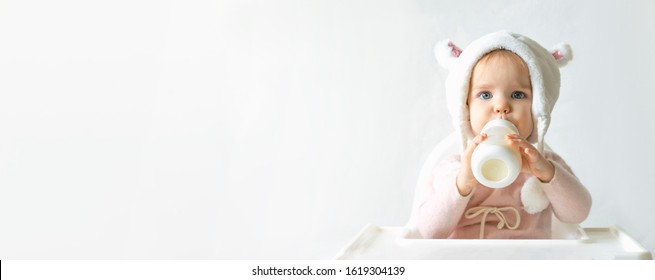 Little toddler girl in a warm fluffy hat drinks milk from a bottle while sitting. Half-length portrait. White gray background. Banner, free space, copy space