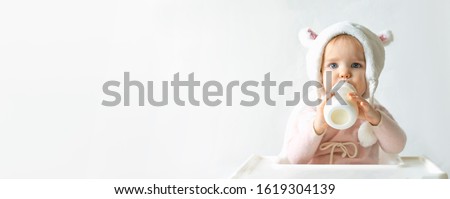 Little toddler girl in a warm fluffy hat drinks milk from a bottle while sitting. Half-length portrait. White gray background. Banner, free space, copy space