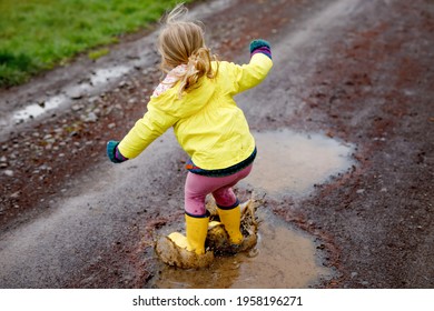 Little Toddler Girl Wearing Yellow Rain Gum Boots, Running And Walking During Sleet. Happy Child In Colorful Clothes Jumping Into Puddle, Splashing With Water, Outdoor Activity. Happiness, Childhood