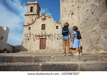 Little travelers do sightseeing in Spain Peniscola old town stand in front of church of the Ermitana point fingers at belltower on sunny hot day, view from behind