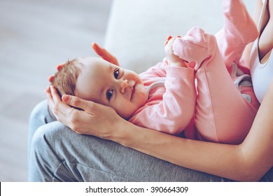 Little Treasure. Close Up Side View Part Of Young Woman Holding Smiling Baby Girl On Her Knees While Sitting On The Couch 