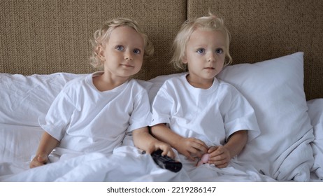 Little Twin Sisters Sit In Bed Leaning On Pillows. They Are Watching TV Which Hangs On The Wall. The Screen Shows Children's Content. From Time To Time They Change Channels.