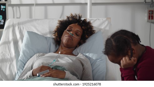 Little Upset African Girl Sitting At Bedside Of Sick Mother In Hospital Ward. Unconscious Afro Woman Patient Lying In Hospital Bed With Little Daughter Visiting Her