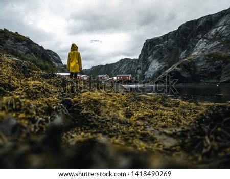 a little village with red houses on the shore and seaweed covered rocks in the foreground with dark water of the ocean and mountains in the background and a girl in a yellow coat standing in the frame