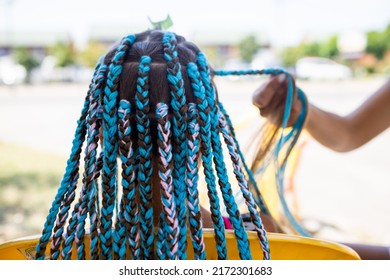 A little waiting tanned girl in a bright yellow summer suit, weaves colored pink-blue African braids in her dark wet thick hair on a hot sunny sea day