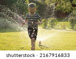 A little wet boy runs barefoot on the lawn next to the sprinkler. Happy carefree childhood and holidays concept. 