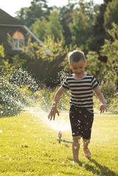 A Little Wet Boy Runs Barefoot On The Lawn Next To The Sprinkler. Happy Carefree Childhood And Holidays Concept. 