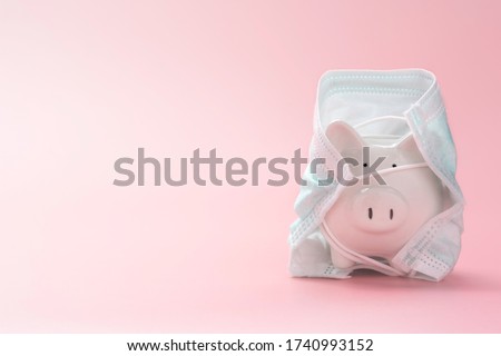 a little white piggy bank, wearing a big surgical face mask or green doctor mask, isolated on pink rose gold background with text space. Money saving concept in time of coronavirus pandemic crisis