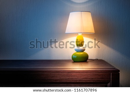 Little yellow, gray and green lamp on a wooden dresser or a night table, illuminating the wall covered with wallpaper at night or late evening. Soft and warm interior mood.