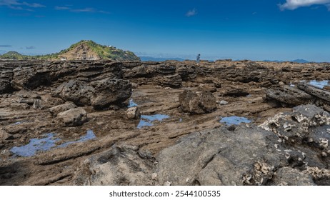 Littoral at low tide. Attached mollusks are visible on exposed rocks. Puddles of water among the boulders. A tiny silhouette of a man walking in the distance. Hill against the blue sky, clouds. - Powered by Shutterstock