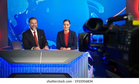 Live News Studio With Beautiful Female And Handsome Man Anchors Start Reporting. TV Broadcasting Channel With Presenters Talking. Television Newsroom Set. Behind The Scene Camera Shooting Shot