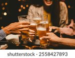 A lively group of young friends celebrates, clinking glasses of beer together in a joyful manner. They chat and smile happily amidst the bustling nightlife and camaraderie.