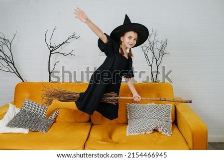 Lively little girl dressed as a witch sitting on a broom, waving her hand. The color of her socks matching the color of the couch that she's standing on, giving the impression that she's flying.