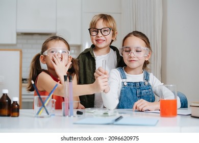 Lively Little Kids Doing Home Science Project, Passing A Flask. All Wearing Glasses. Chemical Glassware And Colored Liquids On The Table.