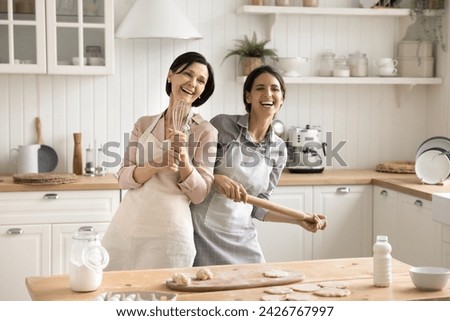 Lively older female, her daughter in aprons cooking together, hold kitchen appliances sing song, having festive mood, celebrate family holidays having fun on carefree pastime at home. Hobby, cookery