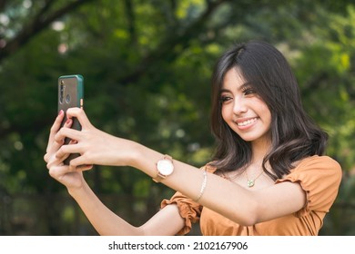 A lively young woman takes a selfie of herself with her phone. Standing outside at a park. - Shutterstock ID 2102167906