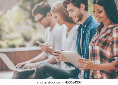 Living In Digital Age. Group Of Happy Young People Holding Different Digital Devices And Smiling While Sitting In A Row Outdoors