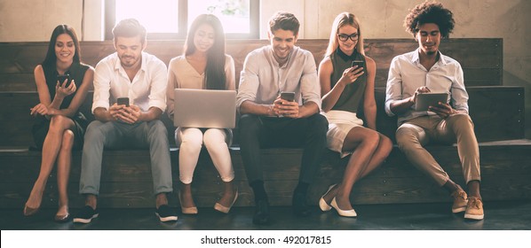 Living In Digitl Age. Front View Of Young People Using Different Gadgets While Sitting Close To Each Other In A Row 