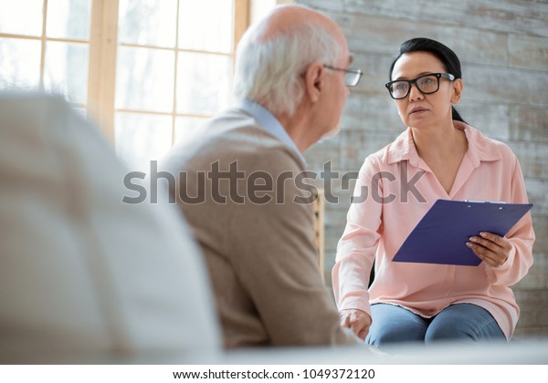 Living environment. Asian\
kind caregiver carrying clipboard while wearing glasses and\
soothing senior man