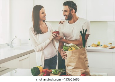 Living a healthy life. Beautiful young couple unpacking shopping bag full of fresh vegetables and smiling while standing in the kitchen together - Powered by Shutterstock