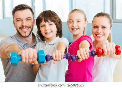 Living A Healthy Life Together. Happy Family Holding Different Sports Equipment While Standing Close To Each Other In Health Club 