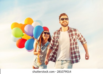 Living Life To The Fullest. Cheerful Young Couple Holding Hands And Smiling While Walking Outdoors With Colorful Balloons