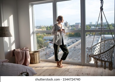 In living room. Blonde woman wearing baggy trousers standing near window in living room