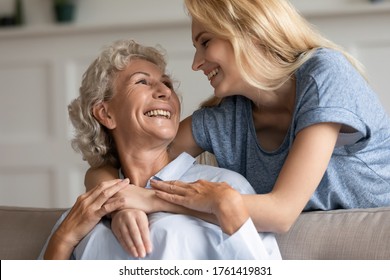 In Living Room Elderly Mom Sit On Sofa While Adult Grown Up Daughter Hugs Her From Behind Relatives Women Looking At Each Other With Warmth Love Close Up. Family Bond, Mother Day Celebration, Concept