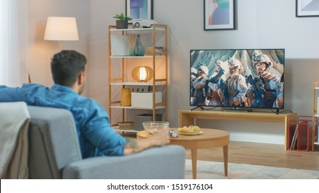 In The Living Room: Guy Relaxing On A Couch Watching War Movie On A TV. Modern Military Warfare Action With War Soldiers Shown On A Television.