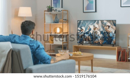 In the Living Room: Guy Relaxing on a Couch Watching War Movie on a TV. Modern Military Warfare Action with War Soldiers Shown on a Television.