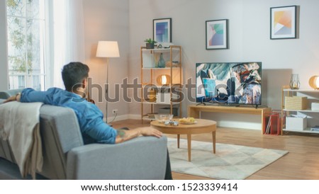 In the Living Room: Guy Relaxing on a Couch Watching War Movie on a TV. Modern Military Warfare Action with War Soldier Shown on a Television.