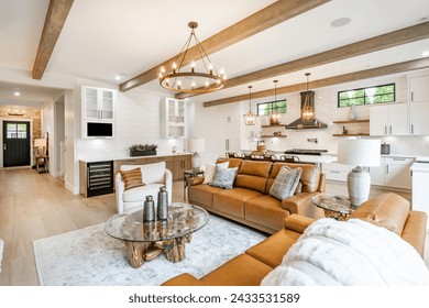 Living room interior with exposed wooden beams on the ceiling carmel colored leather sofa round wagon wheel chandelier fluffy white throw blanket hardwood floor view to kitchen and stone fireplace