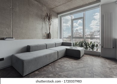 the living room with a long grey sofa near the panoramic window, the walls are decorated as a concrete surface. Minimalist decor, room plants by the window - Shutterstock ID 1720337983