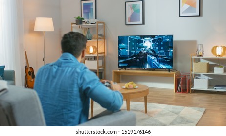 In The Living Room Man Sitting On A Couch Holds Controller Playing In A Console Video Game, 3D Action Shooter Gameplay Shown On TV Screen.