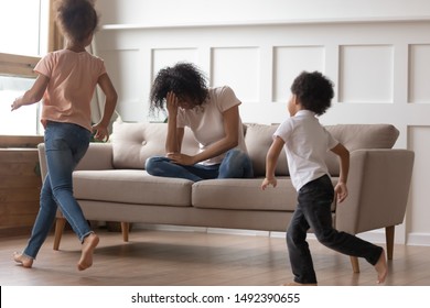 In Living Room Mixed-race Mother Sit On Couch Cover Face With Hand Feels Desperate Unhappy Can't Handle With Noisy Kids Running Near And Make Noise, Misbehave Children, Exhausted Mom Need Rest Concept