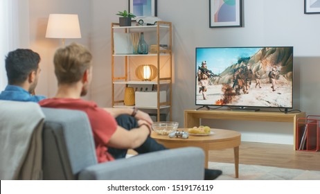 In The Living Room: Two Friends Relaxing On A Couch Watching War Movie On A TV. Modern Military Warfare Action With Shooting And Explosions Shown On A Television.