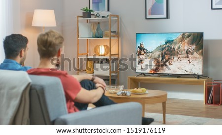 In the Living Room: Two Friends Relaxing on a Couch Watching War Movie on a TV. Modern Military Warfare Action with Shooting and Explosions Shown on a Television.