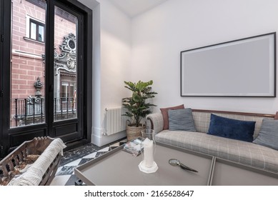 Living room with upholstered sofa with wooden arms, checkerboard marble floors, white armchairs and balcony with aluminum doors overlooking an old building - Shutterstock ID 2165628647