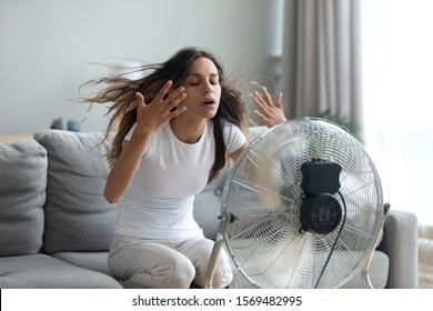In living room without air-conditioner tired from summer heat young woman turned on floor ventilator waving her hands to cool herself, female sitting on couch suffers from unbearable too hot weather - Shutterstock ID 1569482995