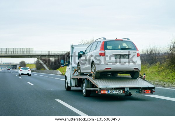 Ljubljana, Slovenia - January 16, 2019: Tow truck\
with car on warranty on road. Trailer and crash auto delivery.\
Vehicles hauler on driveway. European transport logistics. Heavy\
haul trailer