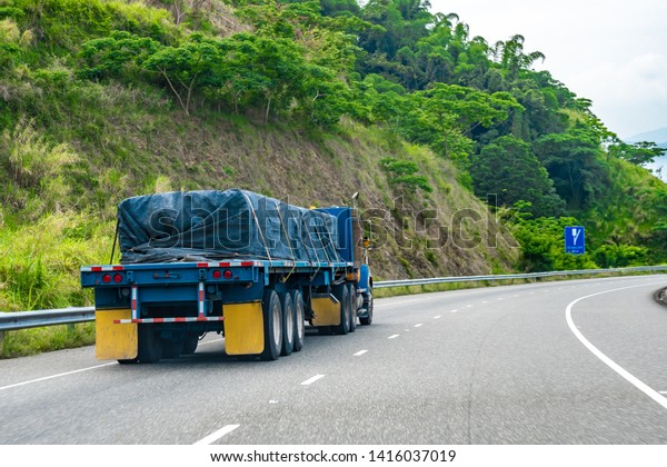 Loaded 22 wheeler cargo semi trailer truck covered\
and tied/strapped down with rope driving on left hand side of road\
on dual carriageway. Red and white reflective stickers/ reflectors\
on rear end.