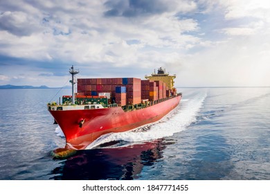 A loaded, red cargo container ship traveling over calm, blue sea with full speed