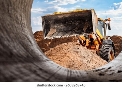 A loader is actively digging and moving a mound of dirt at a construction site, preparing for pipe installation. The bright sky adds to the scene's vitality.