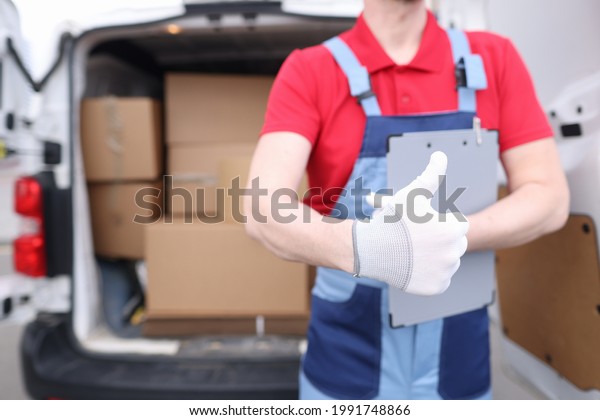 Loader driver in uniform holds a thumbs up in\
background of car with\
boxes