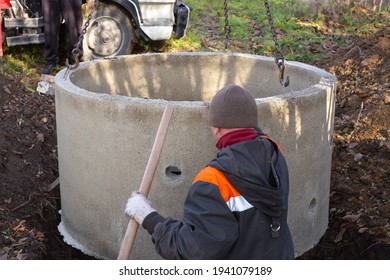 A Loader Lowers A Concrete Ring Into A Dug Hole To Build A Septic Tank. A Worker Installs A Sewer Into The Ground.