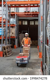 Loader In Workwear Pushing Cart With Goods While Walking Along Racks